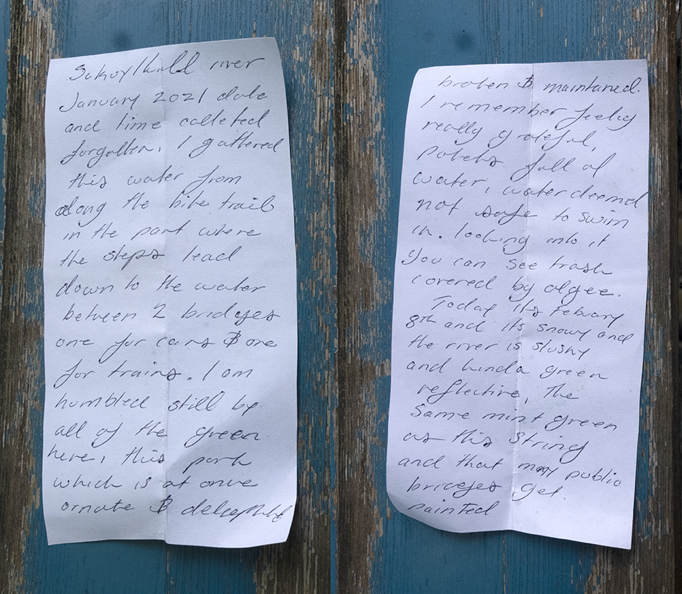 A handwritten note is photographed against wood with peeling blue paint. The note is flipped so both sides are legible, and reads: Schuylkill River January 2021 Exact date and time collected forgotten.  I gathered this water from along the bike trail in the part where the steps lead down to the water, between two bridges, one for cars and one for trains. I am humbled still by all the green here, this park which is at once ornate and dilapidated, broken and maintained. I remember feeling really grateful, potholes full of water, water deemed not safe to swim in. Looking into it, you can see trash covered by algae.   Today it’s February 8 and it’s snowy and the river is slushy and kinda green reflective, the same mint green as this string and that many public bridges get painted.