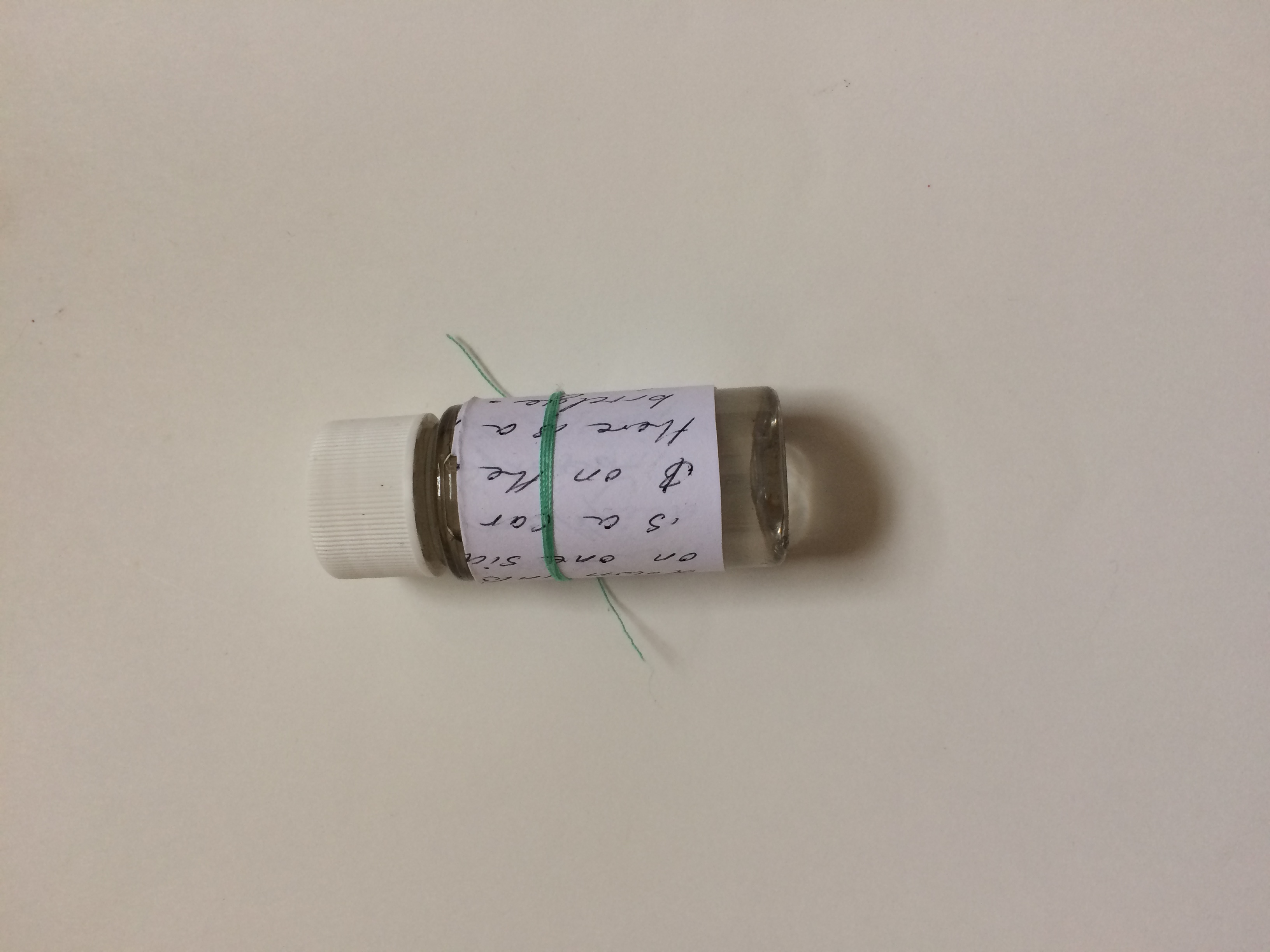 A tiny glass vial of water with a white plastic cap is photographed against a white background. The vial has a narrow roll of paper with illegible handwriting wrapped around it and tied on with a mint green thread.