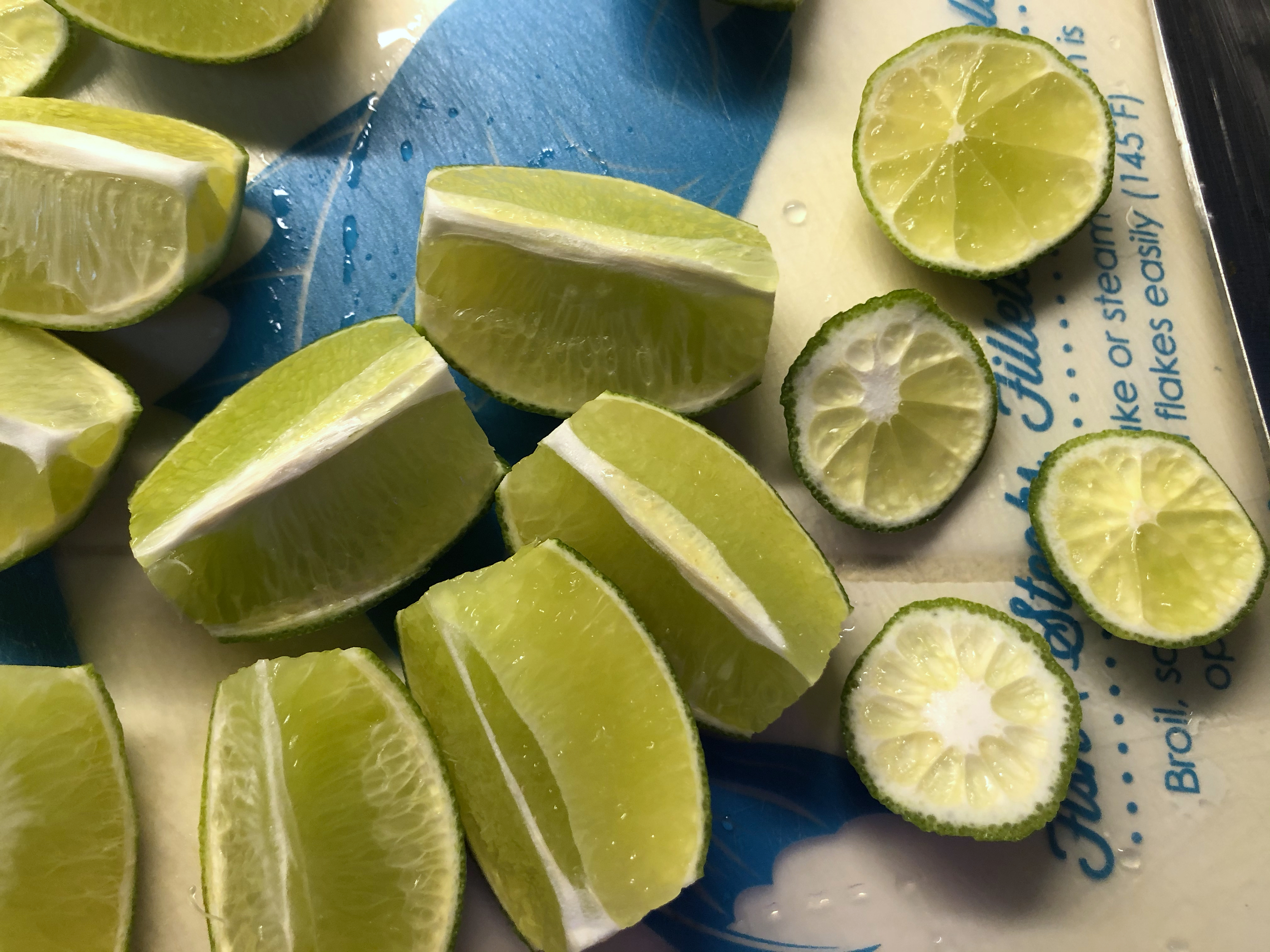 An overhead view of several limes cut into halves and quarters. The limes are placed atop a vintage-looking blue and cream package reading "Fisk Steaks Fillets", with other text obscured.