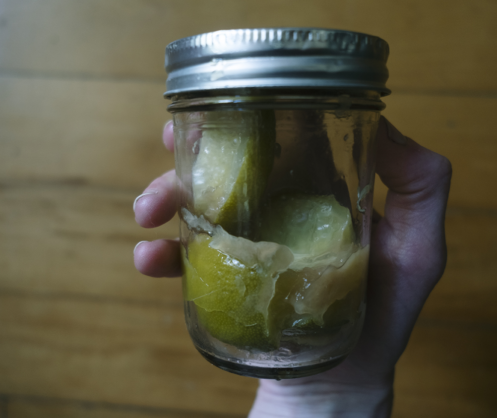 Looking down at one hand holding a glass jar half-full of pickled limes and brine, above a wood floor. 